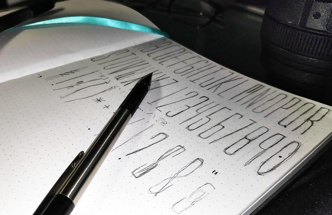 A mechanical pencil over a sketchbook of a font sketched out by Joe Abellard - personal project bio
